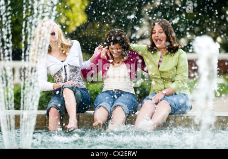 Three girls sit on the edge of a fountain laughing as they dip their feet in the water. Stock Photo