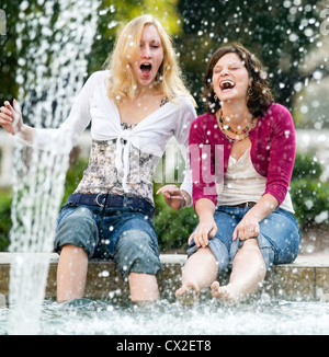 Two girls sit on the edge of a fountain laughing as they dip their feet in the water. Stock Photo