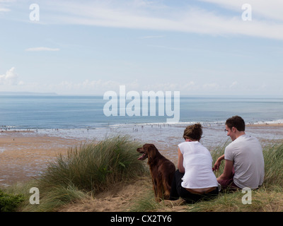 Couple with dog relaxing on sand dunes overlooking Croyde Bay, North Devon. Stock Photo