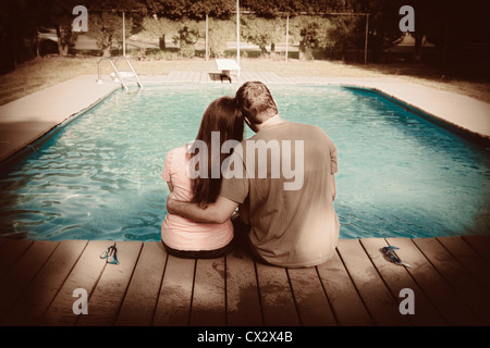 Couple sit on the deck of an in ground swimming pool hugging. Stock Photo