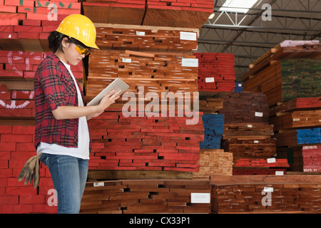 Side view of an Asian female industrial worker working on tablet PC with stacked wooden planks in background Stock Photo