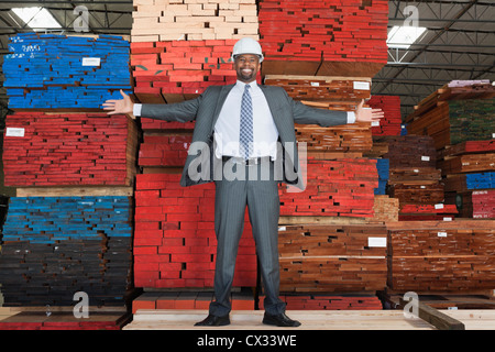 Portrait of happy African American male engineer standing with arms outstretched in front of stacked wooden planks Stock Photo