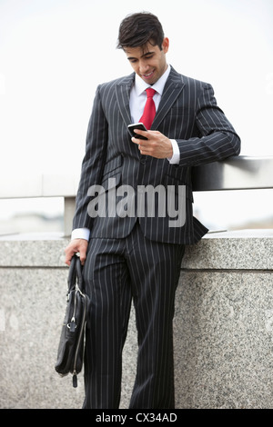 Young Indian businessman texting through cell phone while holding laptop bag at parapet Stock Photo