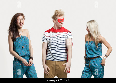 Young man in superhero costume standing with happy female friends in matching jump suits against gray background Stock Photo