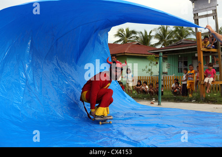 Crazy bearded man in red superhero costume performing a tarp surfing demonstration for local villagers on a skateboard. Stock Photo