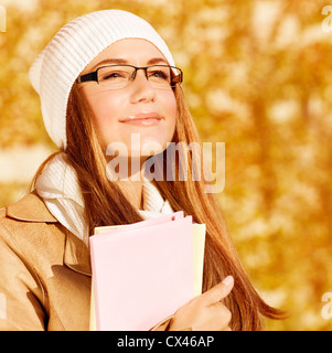 Picture of romantic student girl with textbook on yellow foliage background, closeup portrait of smart female wearing warm hat Stock Photo