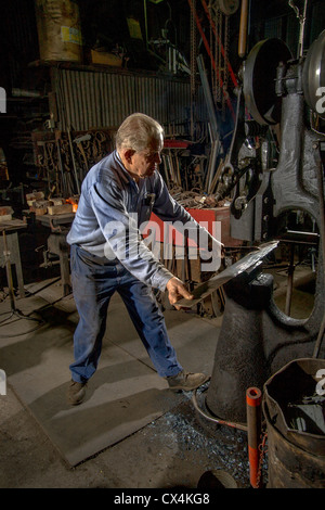 An elderly blacksmith in a Tustin, CA, barn built in 1912 forms a hot saw blade using a trip hammer. Stock Photo