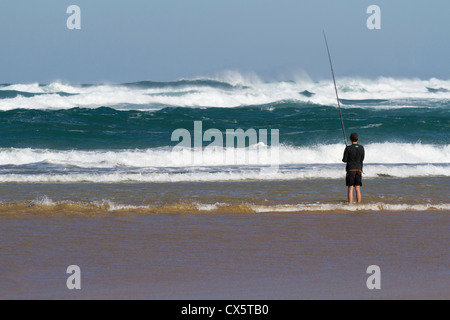 young man fishing from the beach in black shorts and tracksuit top looking away to rough choppy sea's with blue sky Stock Photo