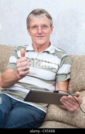 It is a good thing a tablet computer for active senior man. Mature adult showing thumb with tablet PC, sitting on sofa in room Stock Photo