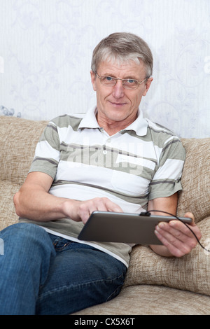 It is a good thing a tablet PC for active senior man. Senior adult Caucasian man interested with a new device, sitting on sofa Stock Photo