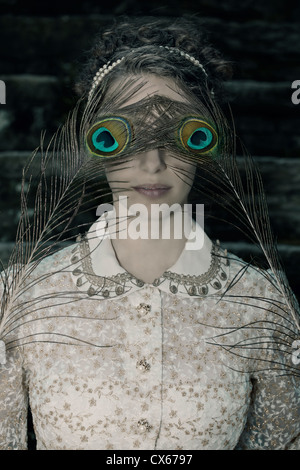 a woman with a victorian dress is holding two peacock feathers in front of her eyes Stock Photo