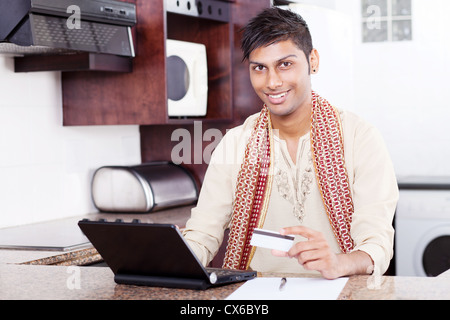 young handsome Indian man holding credit card while using laptop computer at home Stock Photo