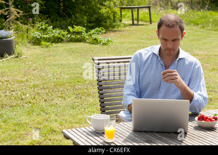 A man sitting at a table in his backyard having breakfast and using a laptop Stock Photo