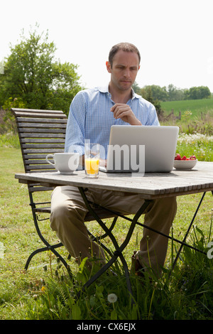 A man sitting at a table in his backyard having breakfast and using a laptop Stock Photo