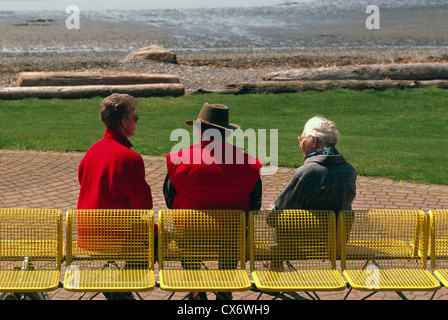 Three Senior People / Friends sitting on a Bench and talking, along a Seaside Promenade Walkway and Waterfront Park Stock Photo