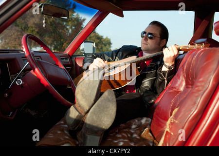 A rockabilly guy playing an acoustic guitar while sitting in his vintage car Stock Photo