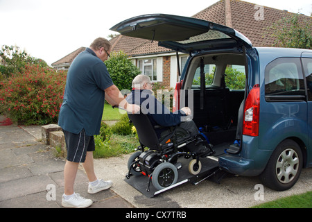 Male Carer son pushing a disabled elderly man in a wheelchair onto a built in ramp in a specially adapted car for getting about