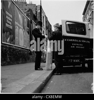 London,1950s. Policeman talks to a parcel delivery man outside his van, while checking his watch to see how long he will give him to leave it at the roadside while he makes his delivery. Stock Photo