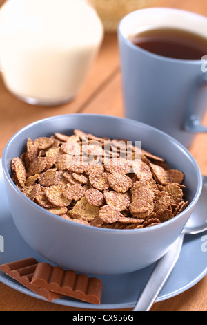 Bowl of chocolate corn flakes cereal with spoon and chocolate bars, a cup of tea and a jug of milk in the back Stock Photo