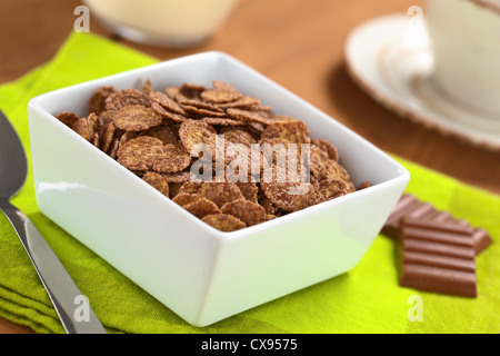 Bowl of chocolate corn flakes cereal with spoon and chocolate bars, a cup and milk in the back Stock Photo