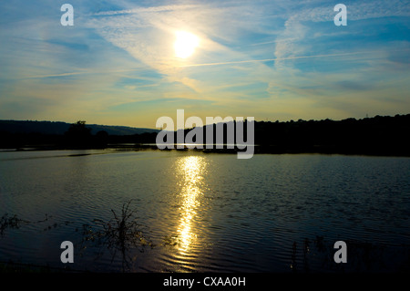 The sun sets over a lake beneath a vapour trail streaked sky, and creates a shaft of sunshine across the water. Stock Photo