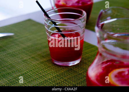 A stylish, ringed glass containing a red coloured punch and a straw stands on a green woven mat between two half full jugs. Stock Photo