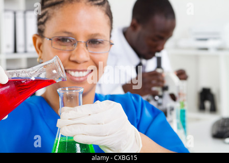 african american female scientist doing experiment in lab, background is her colleague working on microscope Stock Photo