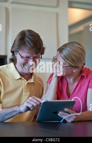 Attractive mature couple at home together relaxing listening to their iPad tablet computer,in light contemporary living room Stock Photo