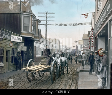 Colorized view of a street scene in Dawson City, Yukon Territory, Canada, 1903. A horse-drawn cart carries lumber on the muddy street and men stand outside various businesses, including John J. Sesnon Company. A banner reading ‘SS Ohio Sails for Seattle Today’ hangs over the street. (Photo by Burton Holmes) Stock Photo