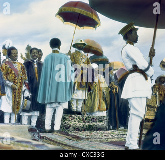 Colorized photo of Ethiopian Emperor Haile Selassie I, born Lij Tafari Makonnen, (1892 - 1975) in the royal enclosure during his coronation, Addis Ababa, Ethiopia, 1930. A man holds an umbrella over Selassie’s head. (Photo by Burton Holmes) Stock Photo