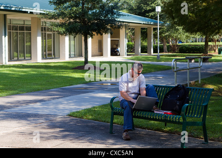 Middle aged student works on his laptop while sitting on a bench on a university campus outdoor area. Stock Photo