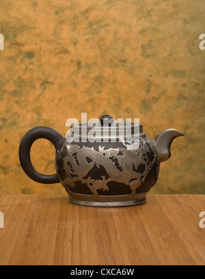 A Chinese teapot with dragon motif made of black clay and pewter, circa 1900. Bamboo table with mango leaf paper background. Stock Photo