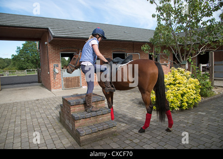 Woman using a mounting block in stable yard to get on to her horse Stock Photo