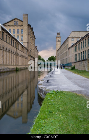 4 people walk on towpath of Leeds Liverpool canal by historic, impressive Salts Mill (buildings reflected in water) - Saltaire, Yorkshire, England, UK Stock Photo