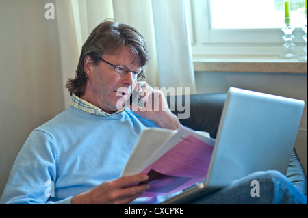 Mature man at home checking business paperwork using his laptop computer and smartphone iPhone 4s Stock Photo