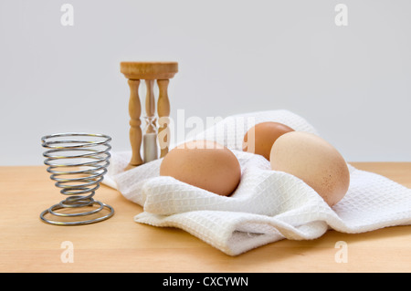 Still life image of egg timer, egg sand egg cup on napkin on top of wooden chopping board against a white background