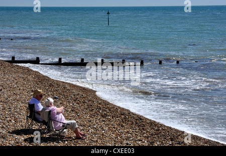 Elderly couple enjoying sea and sunshine - chairs on a pebble beach - gentle waves lapping the shore Stock Photo