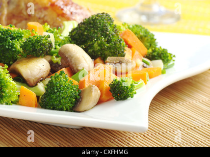 Fried vegetables (broccoli, mushroom, carrot, shallot) on white plate with chicken meat and wine glass in the background Stock Photo