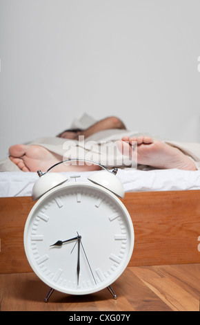 A young tired man laying on a bed. Big alarm clock is placed in front of the bed