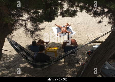 Three people in the shade of the sun whilst a fourth lies on the beach Stock Photo