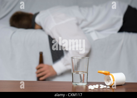 Glass of water and pills on the table. In the background out of focus a passed out businessman on the sofa. Stock Photo