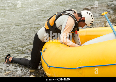 A man wearing a white and beige long-sleeve wetsuit, black wetsuit pants, a white helmet with ventilation slots, and a yellow life vest with black straps is leaning into a bright yellow inflatable Stock Photo