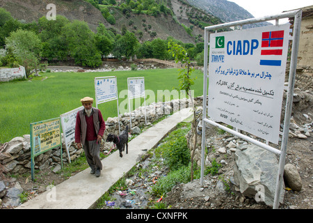 Muslim man leading his goat past signs promoting the projects of various development agencies, Rumbur Valley, Chitral, Khyber-Pakhtunkhwa, Pakistan Stock Photo