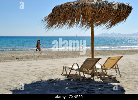 Tourists walking on sandy beach with thatched umbrella and two empty beach chairs view across sea Stock Photo