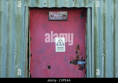 Rusty and faded red door set in corrugated iron wall and padlocked with signs Electric Main Switches and Danger Stock Photo
