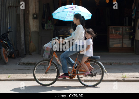 A girl rides on the back seat while her mother pedals her bicycle, holding a parasol. Luang Prabang, Laos. Stock Photo