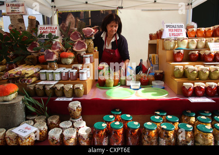 A saleswoman offering typical food products like ham, salamis, pestos, sauces and pickles on the market. Friuli, northern Italy. Stock Photo