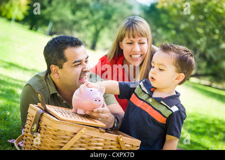 Happy Mixed Race Couple Give Their Son a Piggy Bank at a Picnic in the Park. Stock Photo