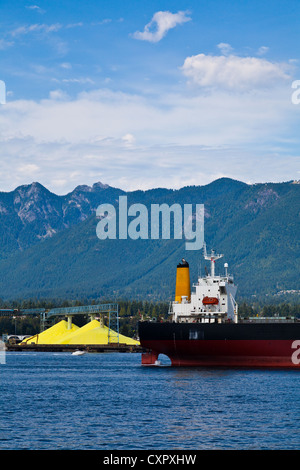 A cargo vessel arriving in Vancouver Harbour, passing piles of processed yellow sulphur. Stock Photo