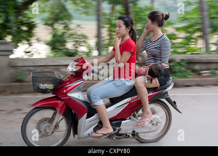 A young woman rides pillion while holding her child across her lap in Luang Prabang, Laos Stock Photo